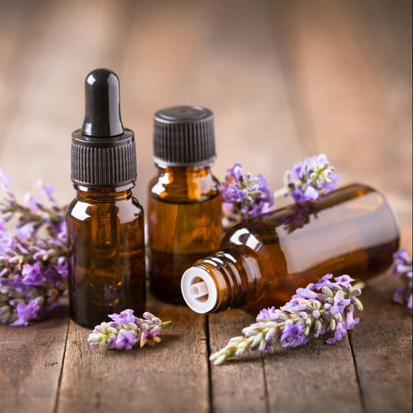 Three small brown glass bottles with droppers on a wooden surface surrounded by lavender flowers.